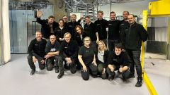 Ein großes, lächelndes Team von Mitarbeitenden in schwarzer Arbeitskleidung posiert gemeinsam in einer Industriehalle vor Produktionsmaschinen.