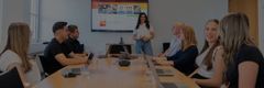 Junge Menschen sitzen an einem Besprechungstisch während eine junge Frau präsentiert