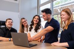 Zwei junge Männer und drei junge Frauen stehen an einem Tisch und schauen auf einen Bildschirm