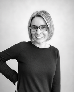 Portrait von Anna Pinkert​, dass sie lächelnd mit kurzen blonden Haaren und Brille vor blauem Hintergrund zeigt.