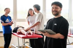 Ein junger Mann lächelt in die Kamera, während im Hintergrund drei Personen eine Patientin auf der Behandlungsliege behandeln.
