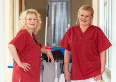 Zwei Reinigungskräfte in roter Arbeitskleidung stehen mit Reinigungswagen vor großen Fenstern.