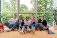 Gruppe junger Leute sitzt lächelnd am Boden und zeigt Daumen hoch.