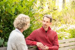 Zwei Menschen sitzen auf einer Bank und unterhalten sich im Garten.