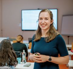 Porträt einer weiblichen Lehrkraft  in einem Unterrichtsraum mit Studierenden, die von hinten zu sehen sind.
