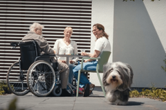 Zwei Mitarbeiterinnen und ein Patient aus dem Hospiz sitzen zusammen am Tisch, vorne weg läuft ein Hund