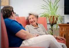 Pflegefachkraft und Bewohnerin sitzen auf der Couch und unterhalten sich freudig