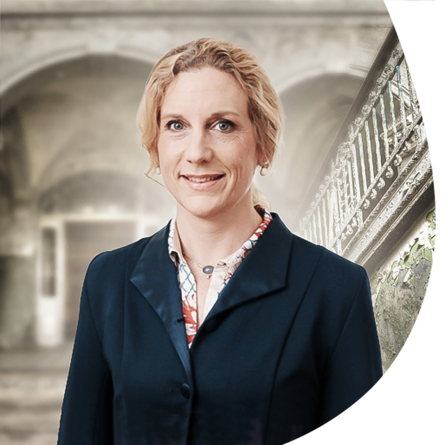 Porträt einer lächelnden Frau in dunklem Blazer vor historischem Klinikambiente mit verwittertem Treppengeländer.