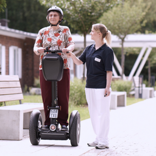 Ältere Patientin auf einem Segway in Begleitung einer Therapeutin beim Mobilitätstraining im Klinikgarten.