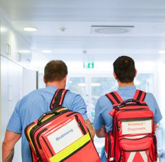 Zu sehen sind zwei Mitarbeiter in Dienstkleidung, bepackt mit Notfallrucksäcken. Sie machen sich gerade auf den Weg zum Einsatz. 
