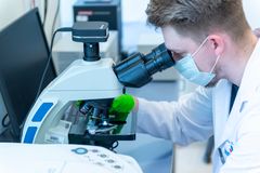 Mężczyzna w fartuchu laboratoryjnym i maseczce patrzy przez mikroskop w laboratorium.