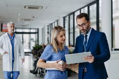 Lekarka w niebieskim uniformie rozmawia z mężczyzną w garniturze który jest Pracownikiem terenowym firmy LEK-AM, oboje patrzą na tablet w szpitalnym korytarzu; w tle widać lekarza w białym fartuchu.