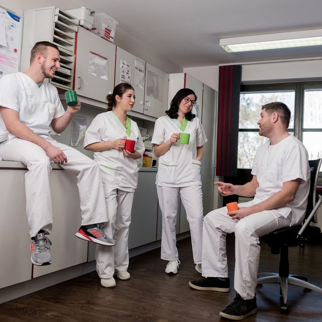 Vier Pfleger:innen in weißer Arbeitskleidung stehen versammelt im Labor, unterhalten sich, machen Pause, lachen und trinken Kaffee.