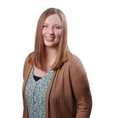 Eine Frau mit langen braunen Haaren lächelt in die Kamera. Sie trägt ein gemustertes Oberteil und eine braune Jacke.