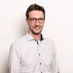 Ein Mann mit dunklen Haaren, Bart und einer Brille. Er trägt ein weißes Hemd und lächelt in die Kamera.