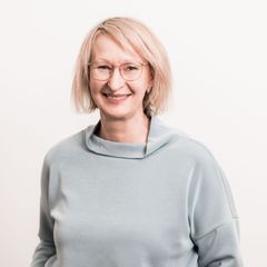 Eine Frau mit blonden, kinnlangen Haaren und Brille. Sie lächelt in die Kamera und trägt ein hellgraues Oberteil.