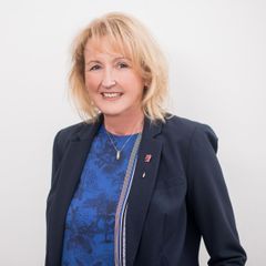 Eine blonde Frau lächelt in die Kamera, trägt einen dunkelblauen Blazer und ein blaues Oberteil darunter.