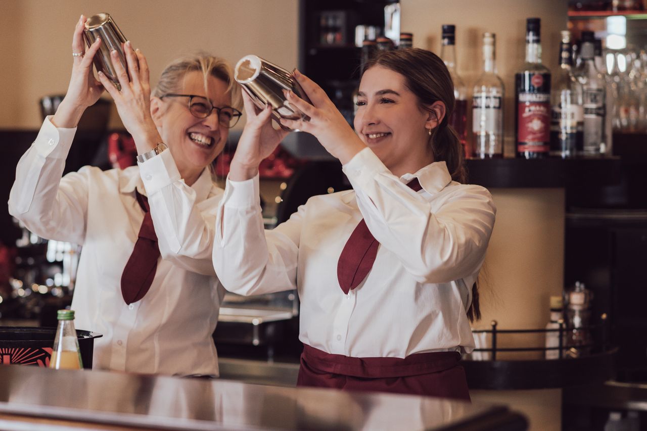Zwei Servicemitarbeiterinnenn stehen an der Bar, mixen ein Getränk und lachen dabei.