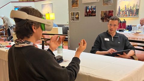 An einem Tisch sitzen sich ein Mitarbeiter und eine Frau, die gerade die VR-Brille testet, gegenüber.