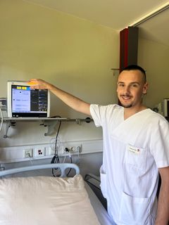 Ein junger Pfleger hat die Hand auf ein technisches Gerät gelegt und blickt lächelnd in die Kamera.