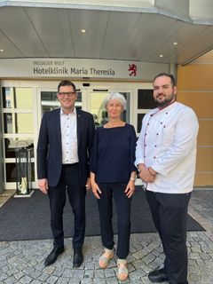 Drei Personen stehen vor der Hotelklinik und blicken lächelnd in die Kamera. Die Person rechts trägt eine Koch-Jacke.