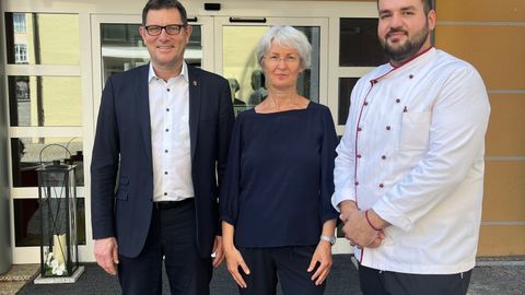 Drei Personen stehen vor der Hotelklinik und blicken lächelnd in die Kamera. Die Person rechts trägt eine Koch-Jacke.