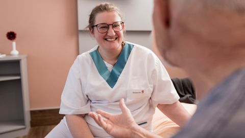 Eine Pflegerin sitzt und führt ein Gespräch mit einem Patienten. Sie lächelt breit.