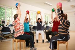 In einem hellen Gymnastikraum mit großen Fenstern sitzen sechs Personen im Kreis auf Holzstühlen. Fünf Teilnehmende heben bunte Gymnastikbälle mit gestreckten Armen über den Kopf. In der Mitte sitzt eine Therapeutin in weißer Hose und dunkelblauem Shirt, die die Bewegungsübung anleitet. Alle Personen sind in sportlicher Kleidung und machen aktiv mit. Im Hintergrund stehen Bürostühle und Trainingsgeräte. Der Raum ist freundlich und funktional eingerichtet.