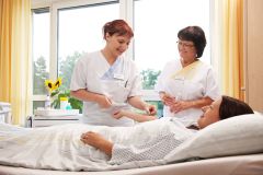 In einem hellen Patientenzimmer mit großen Fenstern und gelben Vorhängen betreuen zwei Pflegekräfte eine Patientin. Die Patientin liegt im Bett und trägt ein Krankenhausnachthemd. Eine Pflegekraft beugt sich lächelnd über sie und hält ihren Arm, während die andere daneben steht und eine kleine Verpackung in den Händen hält. Beide Pflegekräfte tragen weiße Arbeitskleidung. Im Hintergrund stehen medizinisches Gerät und eine Vase mit einer Sonnenblume auf einer Kommode. Draußen sind Bäume zu sehen.