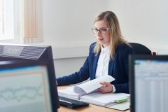 Eine Frau mit langen blonden Haaren und Brille sitzt an einem Schreibtisch in einem Büro. Sie trägt ein dunkelblaues Sakko über einer weißen Bluse und arbeitet an einem Computer. In der einen Hand hält sie Unterlagen, mit der anderen bedient sie die Maus. Im Vordergrund sind zwei Bildschirme mit Tabellen und Grafiken unscharf zu erkennen. Das Büro ist hell und schlicht eingerichtet, Tageslicht fällt durch ein Fenster mit hellem Vorhang.