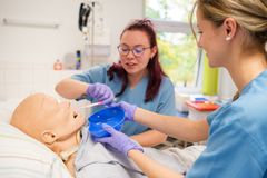 Pflegekräfte im blauen Kittel putzen einer Patientenpuppe im Krankenbett die Zähne.