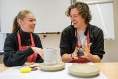 Eine Schülerin und ein Schüler mit dunklen Pullovern und roter Schürze formen Gefäße aus Ton.