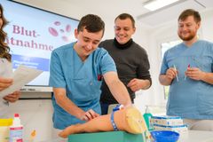 Lächelnde Personen in medizinischer Kleidung üben die Blutentnahme an einem Blutentnahme-Arm.