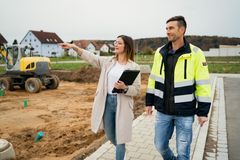 Eine Frau mit Klemmbrett und ein Monteur in Sicherheitsweste tauschen sich vor einer Baustelle über ein Projekt aus