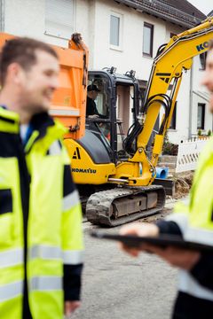 Im Hintergrund ein Bagger der einen Graben für eine Stromleitung aushebt und im Vordergrund zwei Männer in Arbeitskleidung, die den Baustelleneinsatz besprechen.