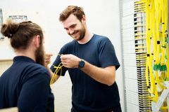 Zwei Männer in blauen T-Shirts stehen vor einem Schaltschrank für Nachrichtennetze und prüfen Steckverbindungen.