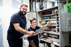 Eine Elektrofachkraft der REWAG mit einem Ordner in der Hand und ein Auszubildender, die zusammen eine Schaltanlage in einem REWAG-BHKW überprüfen.