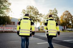Zwei REWAG-Mitarbeiter in gelb-blauen Arbeitsjacken von hinten, die gerade eine Straße hin zu einer Baustelle am Straßenrand überqueren.