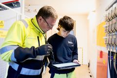 Ein Elektromonteur in gelb/blauer Arbeitssicherheitskleidung und Sicherheitshandschuhen zusammen mit einem Auszubildenden in blauem Sweatshirt und Arbeitshose stehen in einer Trafostation. Der Auszubildende hält ein Tablet in der Hand. Beide schauen hinein und überprüfen etwas.