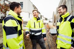 Drei junge Elektrofachkräfte stehen mit gelben Sicherheitswesten in einer Baugrube. Die drei lächelnden Männer tauschen sich über Projekte  aus