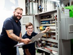 Eine Elektrofachkraft mit einem Ordner und ein Azubi der eine Schaltanlage betätigt. 