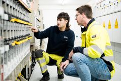Eine Elektrofachkraft und ein Azubi, mit Sicherheitsanzügen, arbeiten gemeinsam an einer Schaltanlage