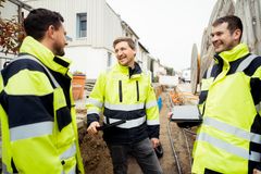 Drei Männer der REWAG in gelben Arbeitsjacken, die in einem Baustellengraben stehen. Zwei haben ein Tablet in der Hand. Sie lächeln. Im Hintergrund ist der Graben mit diversen Kabeln und Rohren, große Kabeltrommeln und ein Einsatzfahrzeug zu sehen.
