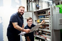 Eine Elektrofachkraft der REWAG mit einem Ordner in der Hand und ein Auszubildender, die zusammen eine Schaltanlage in einem REWAG-BHKW überprüfen.