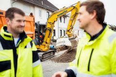 Baggerführer in einem kleinen Bagger, der in einem Wohngebiet gerade einen Graben für eine Stromleitung ausbaggert. Am Rande sind unscharf zwei REWAG-Mitarbeiter in gelber Arbeitskleidung zu sehen.