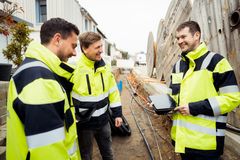 Drei Männer in gelben Arbeitsjacken, die in einem Baustellengraben stehen. Zwei haben ein Tablet in der Hand. Sie lächeln. Im Hintergrund ist der Graben mit diversen Kabeln und Rohren, große Kabeltrommeln und ein Einsatzfahrzeug zu sehen.
