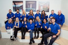 Ein motiviertes Team, mit Daumen nach oben zeigend, an Mitarbeiterinnen und Mitarbeitern der REWAG in ihren blauen Firmenhoodies sitzen in zwei Reihen im Wartebereich des REWAG Kunden-Centers.