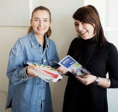 Das Bild zeigt zwei Frauen, die Flyer in den Händen halten und lächelnd in die Kamera  schauen.