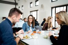 Das Bild zeigt eine lächelnde Frau, die zu einem Mann schaut. Gemeinsam sitzen die beiden mit anderen Kollegen, welche im Hintergrund zu sehen sind, in der Kantine vor ihren mit Essen gefüllten Tellern.