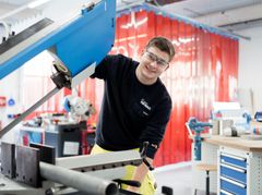 Das Bild zeigt einen lächelnden Mann, der mit Schutzbrille vor einer Maschine steht. Das Bild soll auf die Ausbildung zum Industriemechaniker bei der REWAG hinweisen